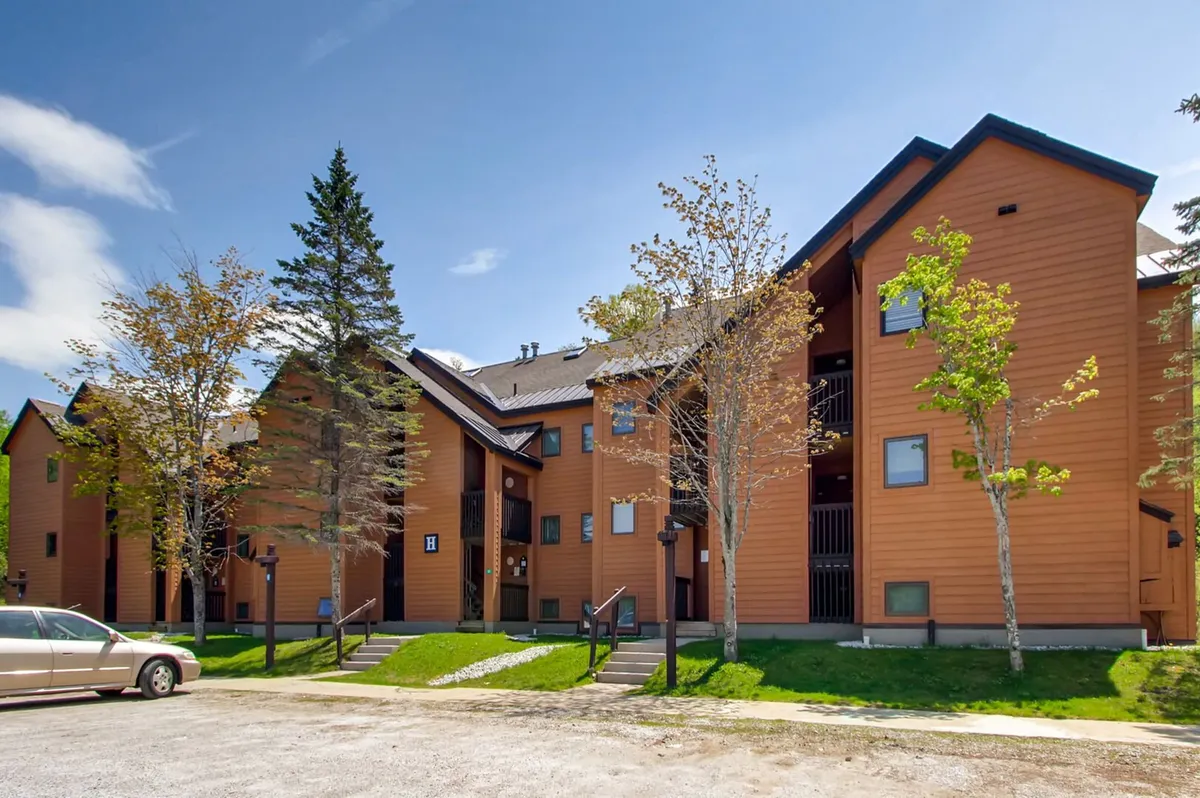 This image shows the inviting wooden exterior of the building surrounded by a landscaped front yard, tall trees, and a pathway leading to the entrance. The natural setting and open pathway suggest accessibility and a welcoming atmosphere, perfect for families arriving for their mountain retreat.