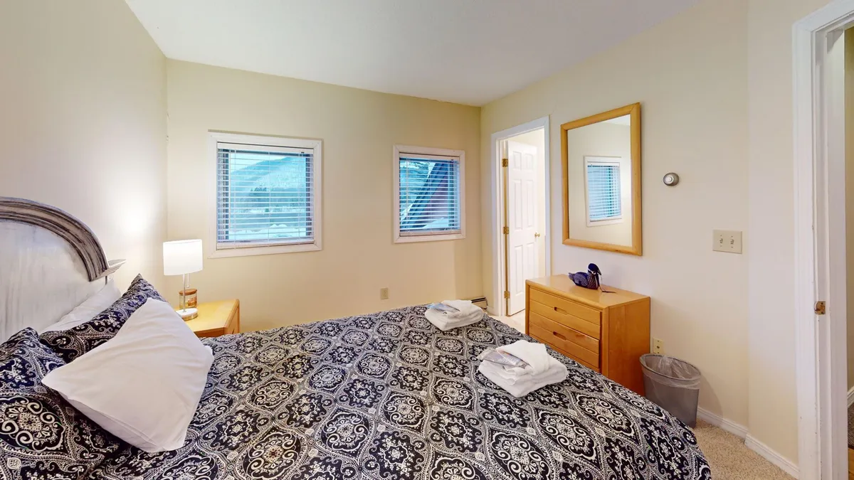 A tranquil bedroom scene featuring a bed with patterned bedding, a nightstand with lamp, wooden dresser, and mirror. The room is softly lit, exuding comfort and a homey vibe ideal for unwinding.
