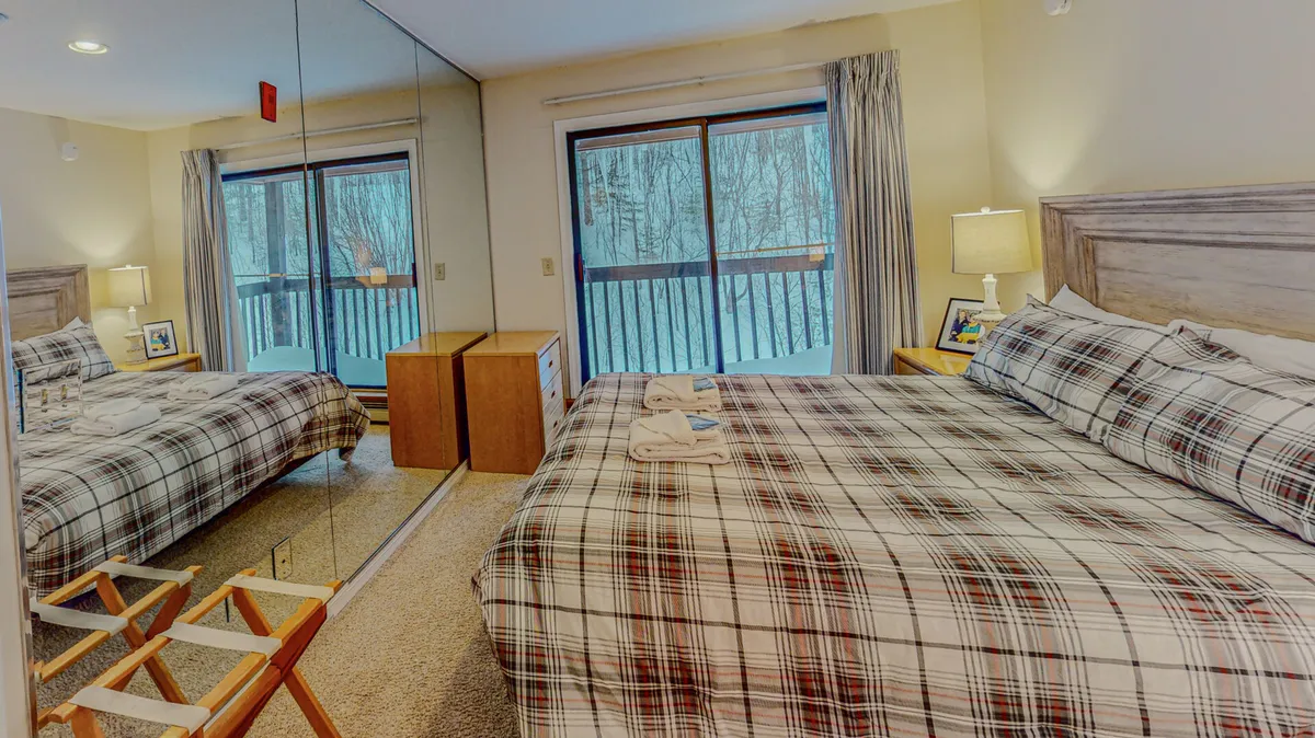 This image shows a bedroom with plaid bedspread, a large mirror, and a balcony view. Soft natural light filters through, highlighting the inviting textures and tranquil ambiance, while the hint of an outdoor balcony connects the interior with the mountain landscape.
