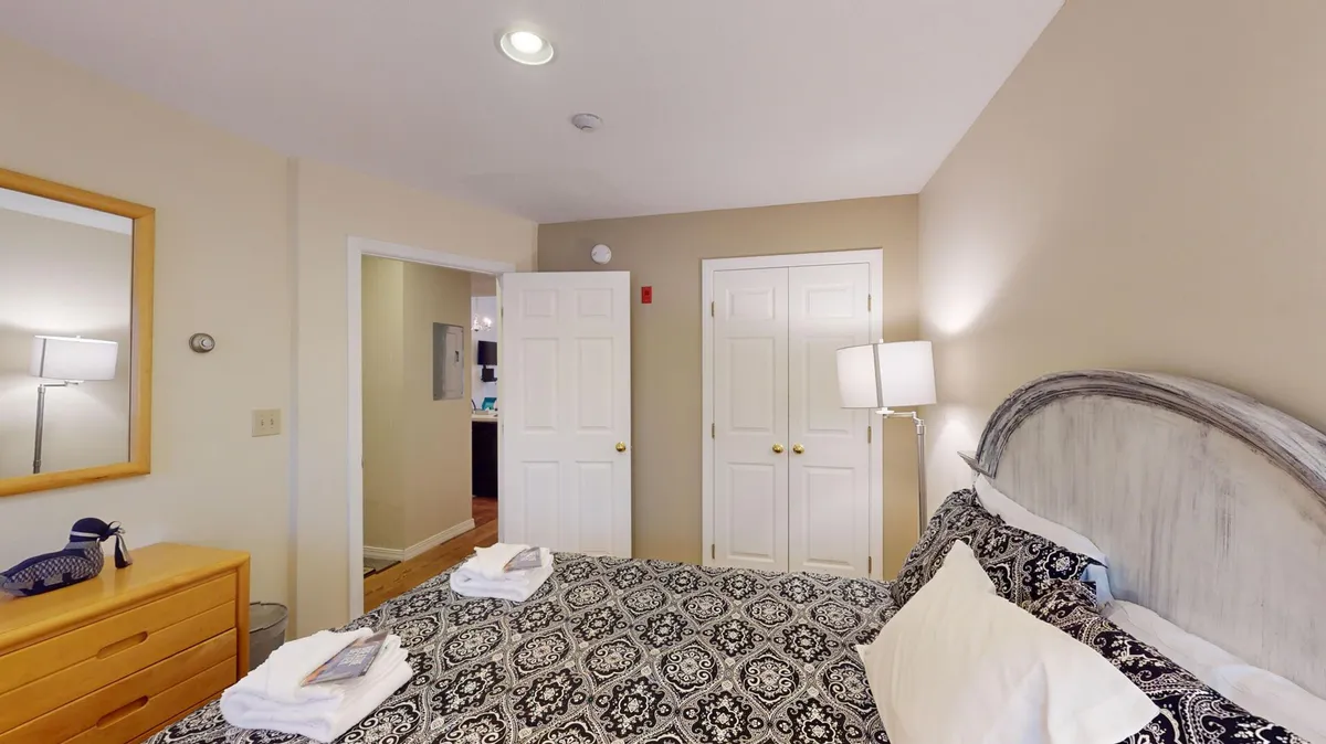 A cozy bedroom with patterned bedding, a mirror, wooden dresser, and a warm lamp on the nightstand. The space is softly illuminated, creating a welcoming environment ideal for winding down after an active day.