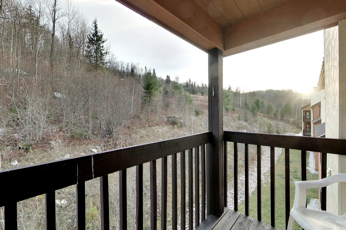 A serene balcony scene with wooden railing overlooking a natural landscape bathed in sunset lighting. The view features distant trees and suggests quiet moments of relaxation while immersed in mountain scenery.