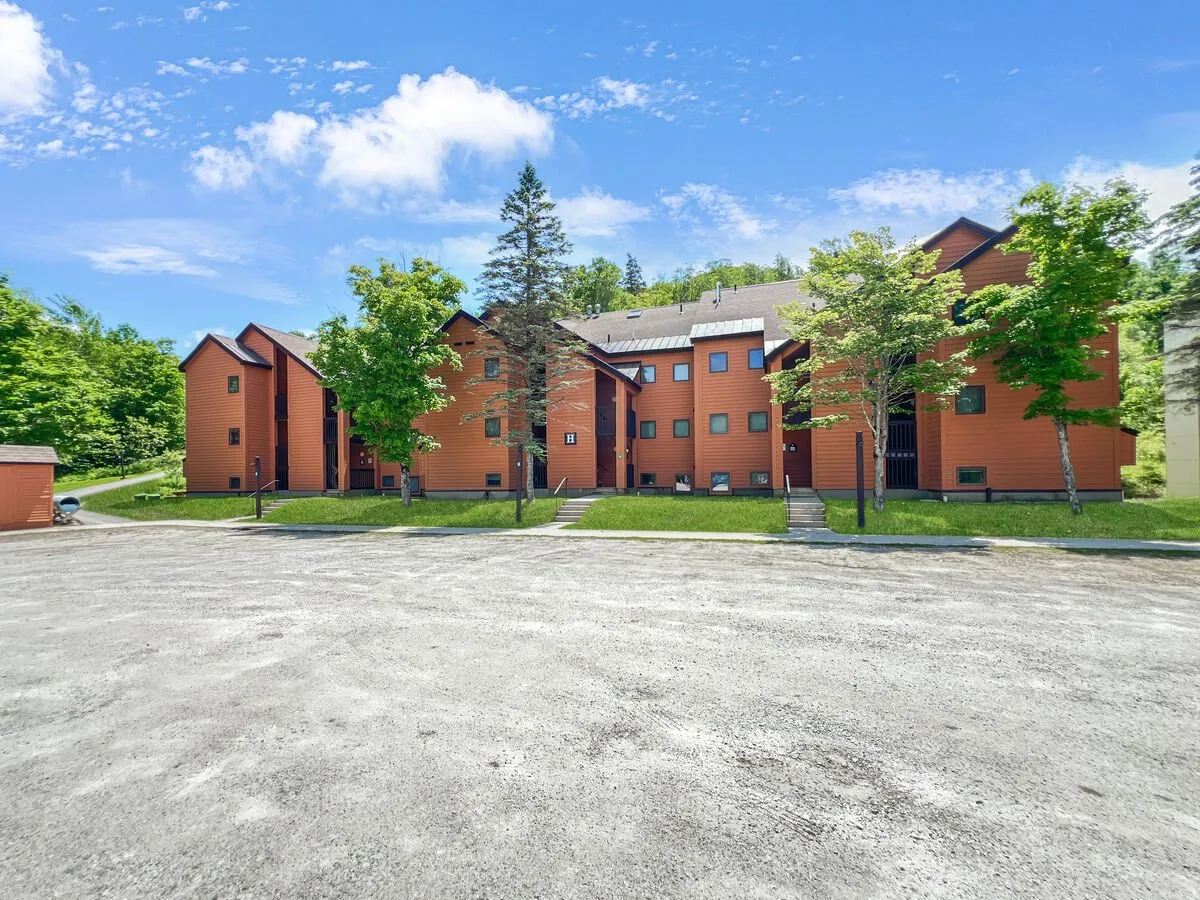 The image captures vibrant orange wooden resort buildings framed by green trees and a clear blue sky. The open, communal space hints at outdoor gatherings, picnics, and nature walks, reinforcing the active, family-friendly spirit of the resort.