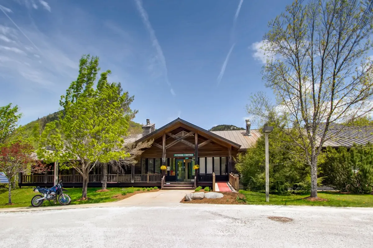 This scene features the wooden entrance to the building, large windows overlooking landscaped gardens, and a stunning mountain backdrop. The setting invites community gatherings, outdoor activities, and nature walks right outside the door.