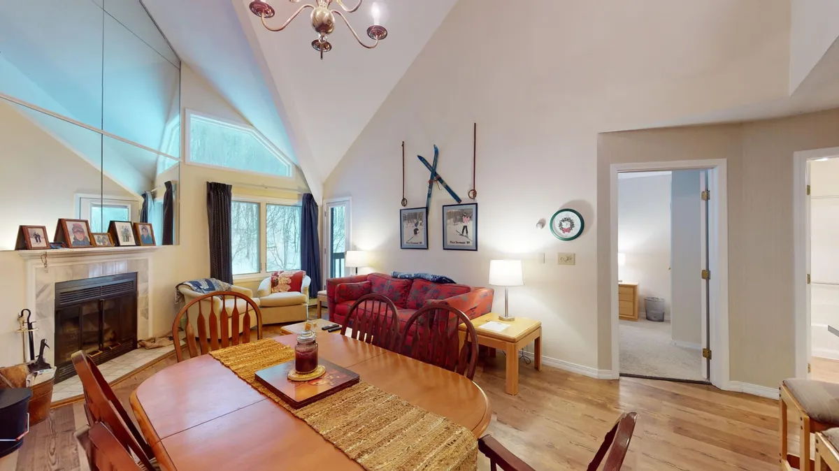 A wide, social living and dining area features a large dining table set for a meal, a plush sofa for lounging, and a crackling fireplace surrounded by artful decor. Expansive windows reveal natural views, hinting at the proximity to the slopes and inviting light into the heart of the home.