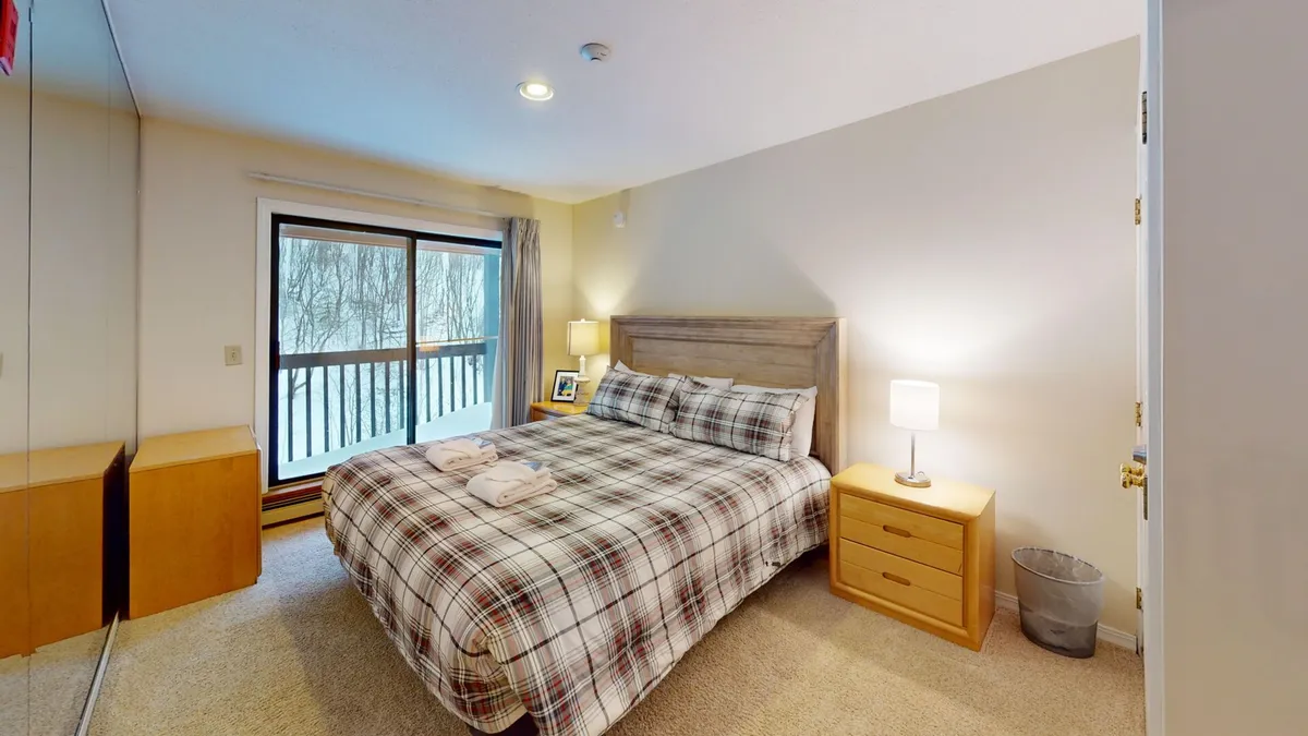A cozy bedroom scene featuring plaid bedding, a nightstand with a lamp, and a large window offering a view of the surrounding forest. The soft natural light and inviting atmosphere suggest moments of relaxation and quiet reflection.